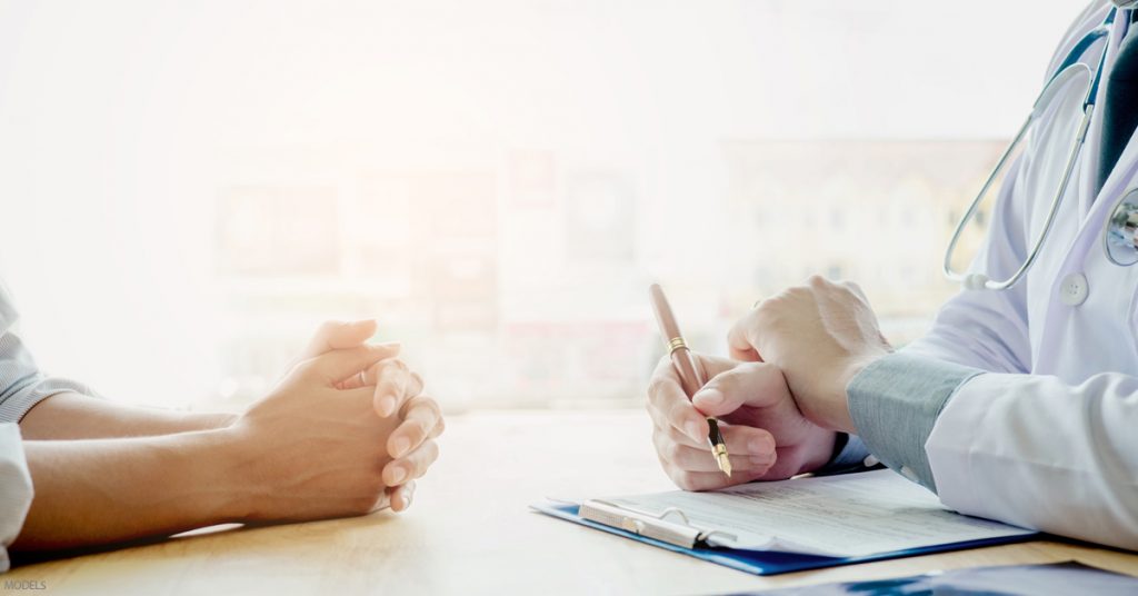 Doctor takes notes with a woman's hands folded on the opposite side of the table.