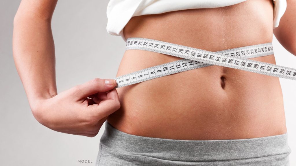 Woman in grey undergarments using a measuring tape around her waist. (MODEL)