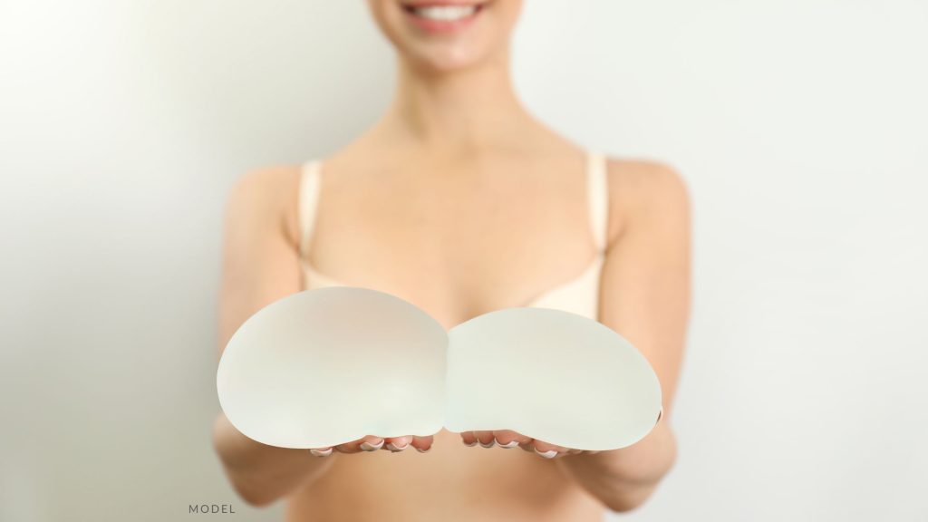 Woman in a white bra holding two implants. (MODEL)