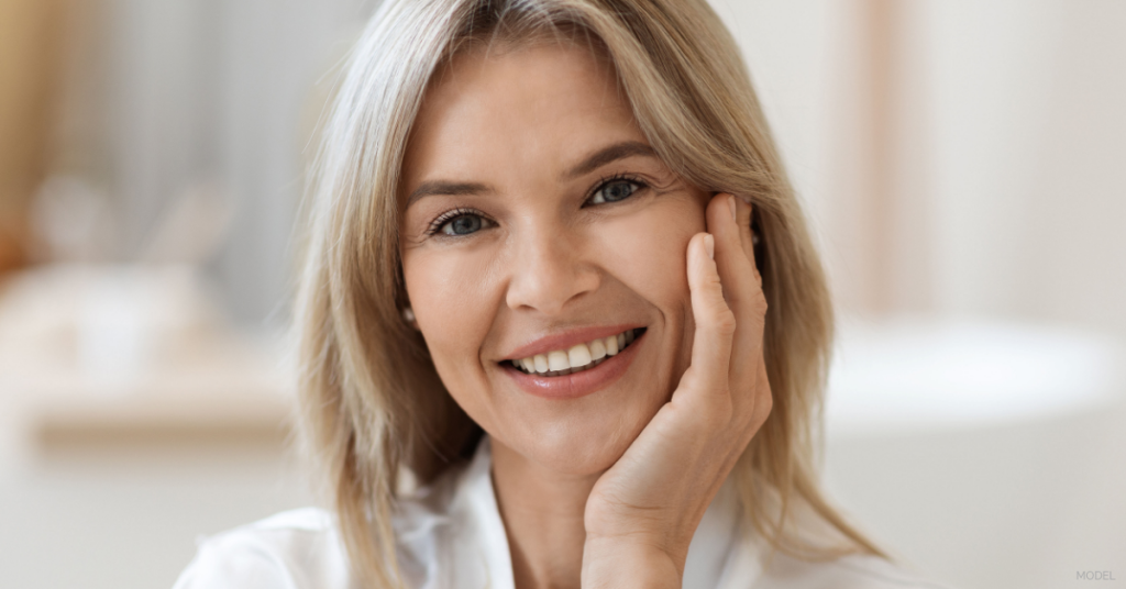 A smiling mature woman gently touching her cheek with one hand (model).